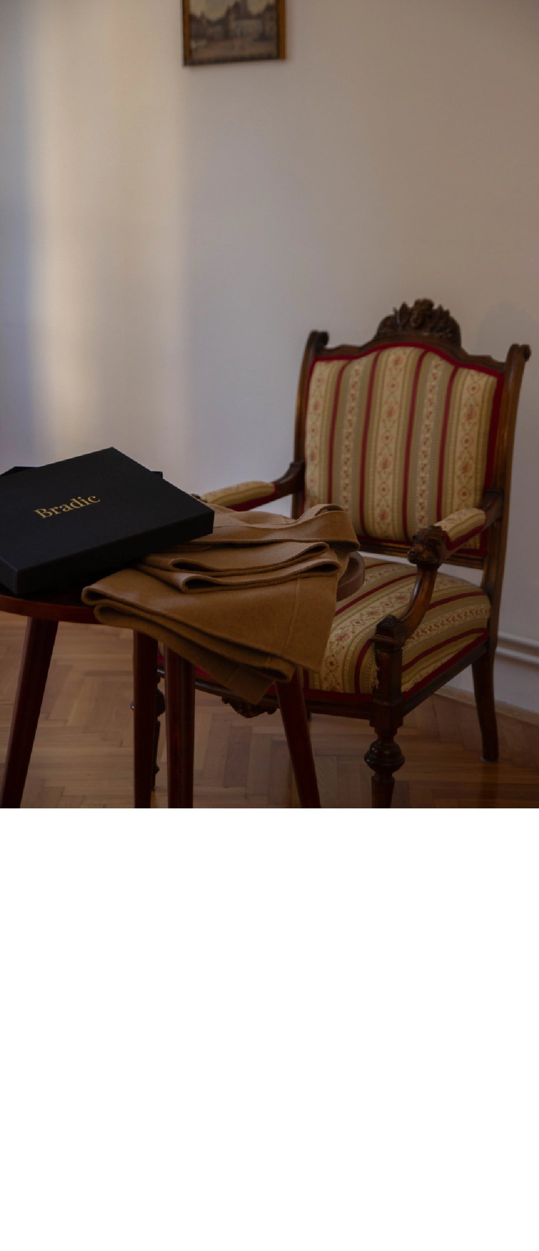 A folded cashmere double faced wrap coat rests on a wooden table next to a black box with the brand name 'Bradic' in gold lettering. The scene is set in a room with a patterned armchair and a light-colored wall.
