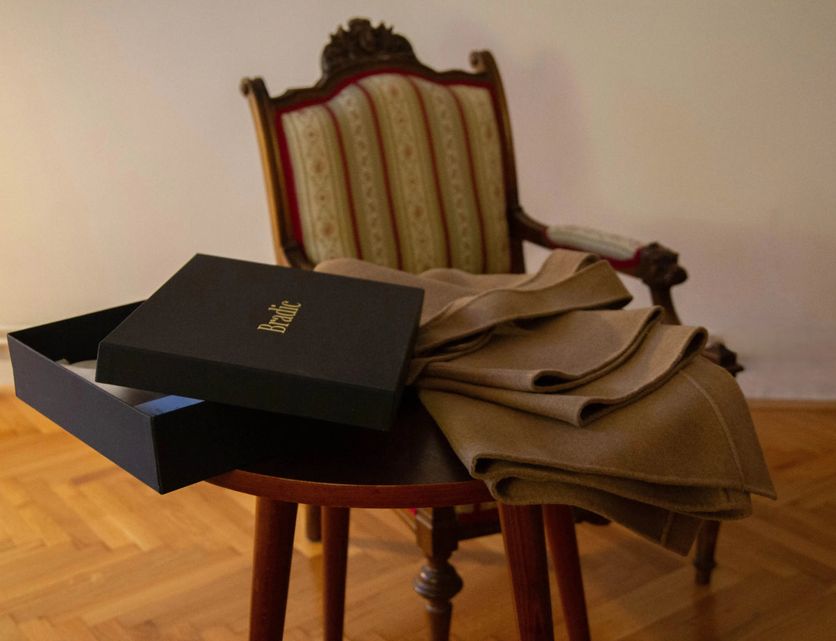 A signature Bradic packaging sits open on a wooden table next to a folded cashmere double faced coat, with an ornate upholstered armchair in the background.
