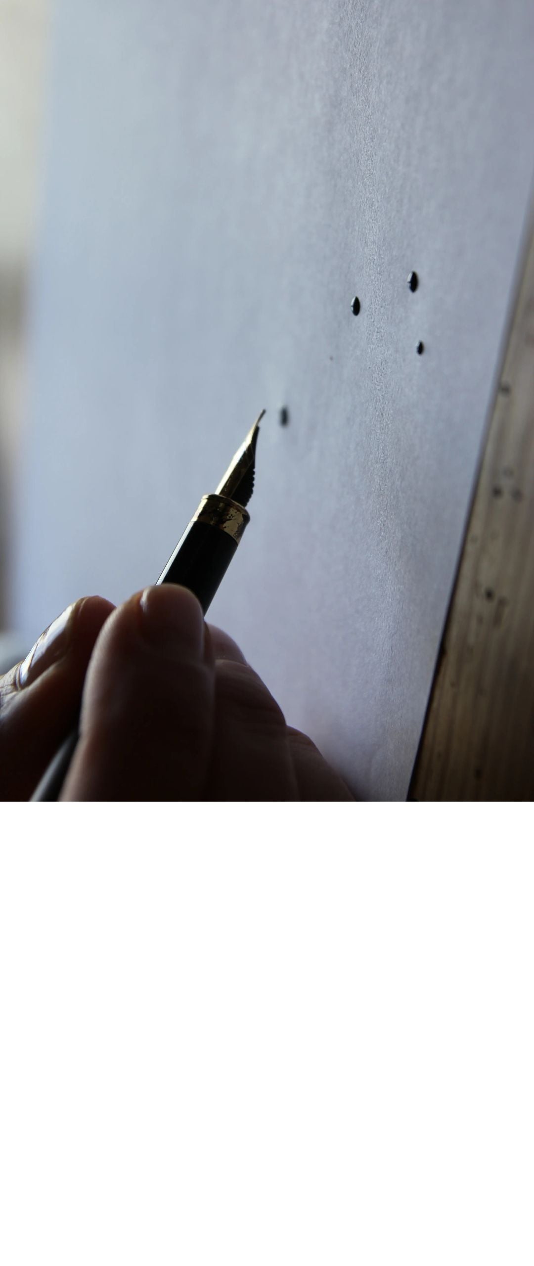 Hand holding a pen over a piece of paper with black ink drops.