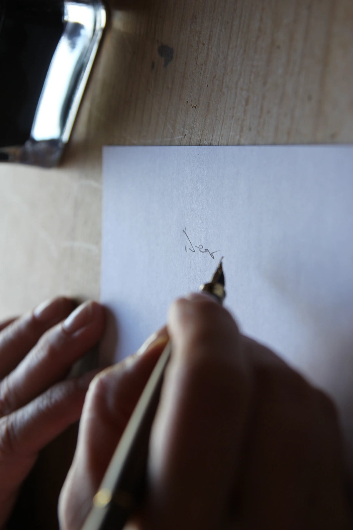 Hand holding a ink dip pen over a piece of paper with writing on a wooden surface