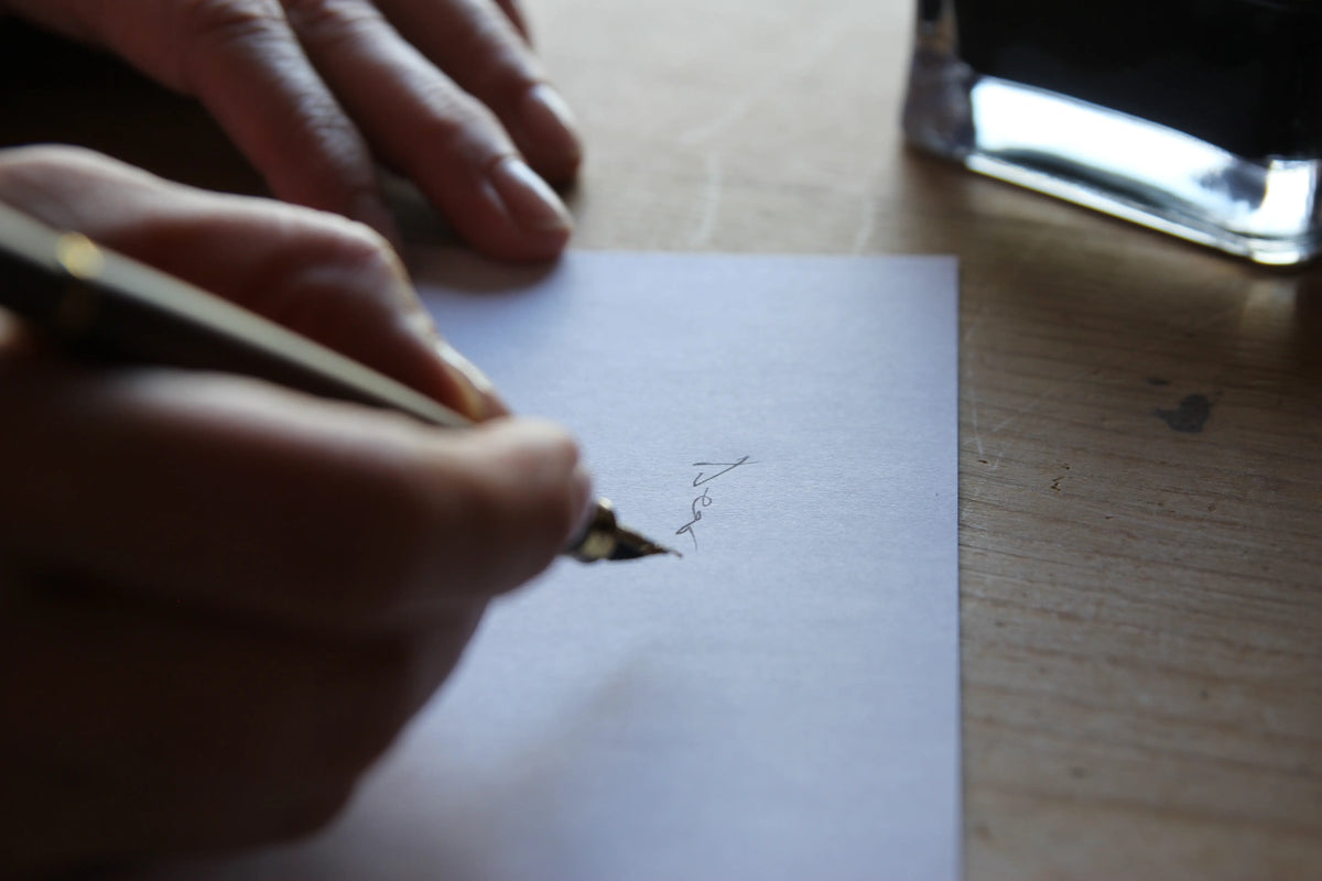 Hand writing on a piece of paper with a ink dip pen on a wooden surface.