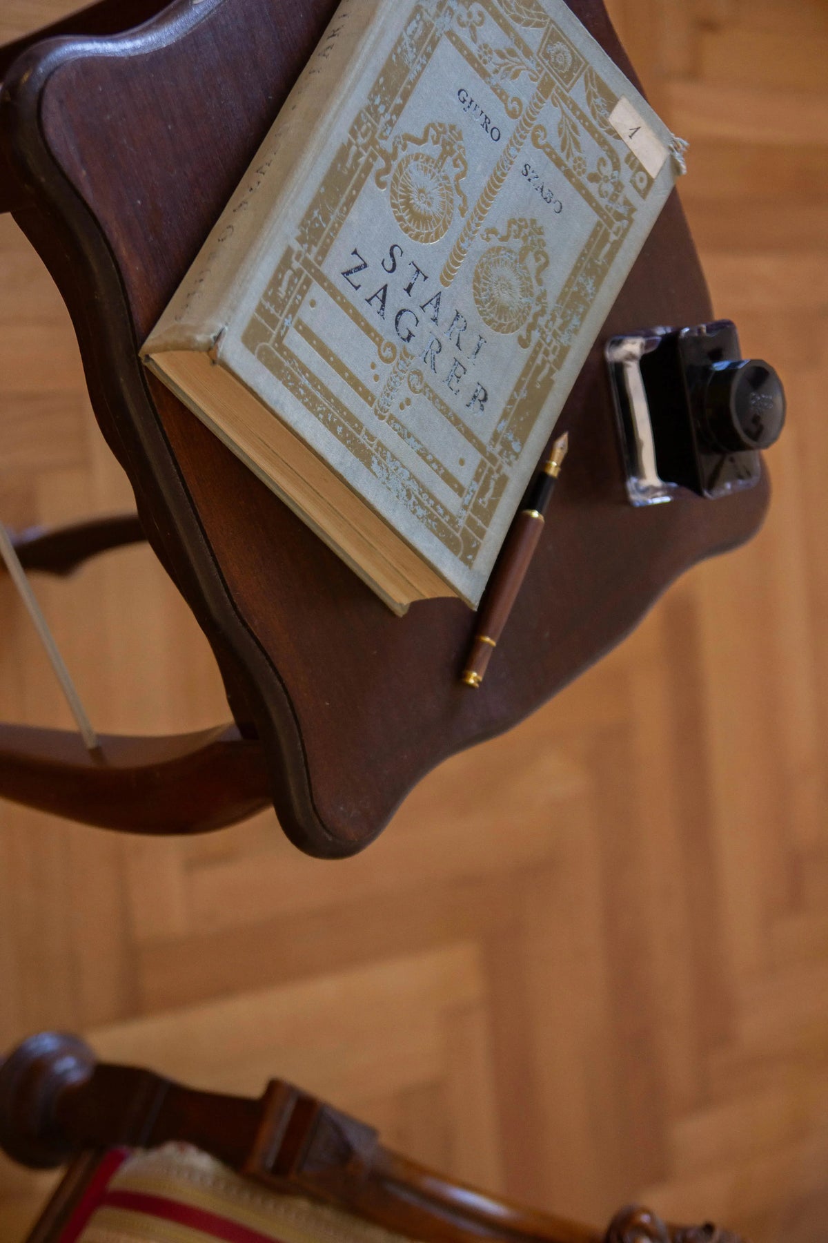 A vintage Zagreb book with ornate gold detailing on a cream cover, resting on a dark wooden table alongside a ink dip pen, set against a warm wood parquet floor.